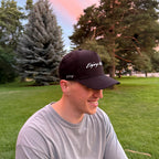 Man wearing a black UPF 50+ cap with Enjoy the Vu logo sitting outdoors on grass with trees in the background