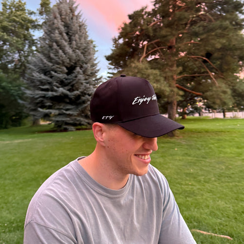Man wearing a black UPF 50+ cap with Enjoy the Vu logo sitting outdoors on grass with trees in the background