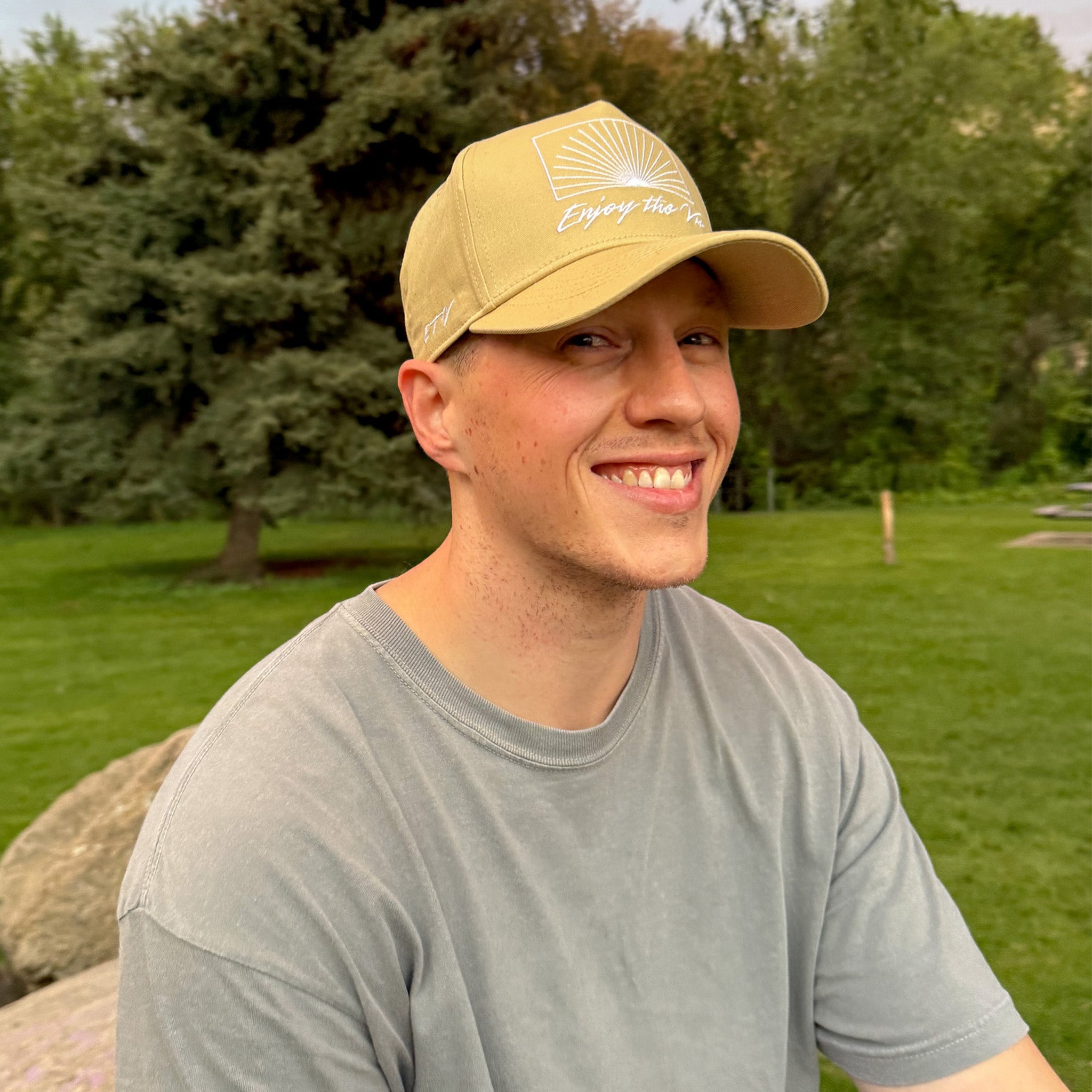 Man wearing a beige UPF 50+ cap with a logo and text, standing outdoors in a park-like setting.