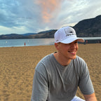 Man wearing a white UPF 50+ cap and gray shirt on a beach with mountains and water in the background