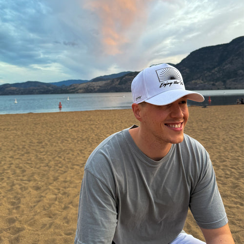 Man wearing a white UPF 50+ cap and gray shirt on a beach with mountains and water in the background